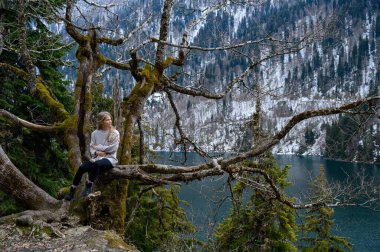 Kız ağaçta oturur ve mavi göl arka planında meditasyon yapar. Abhazya 'daki Ritsa Gölü
