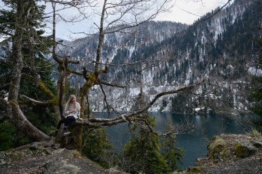 Kız ağaçta oturur ve mavi göl arka planında meditasyon yapar. Abhazya 'daki Ritsa Gölü