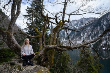Kız ağaçta oturur ve mavi göl arka planında meditasyon yapar. Abhazya 'daki Ritsa Gölü