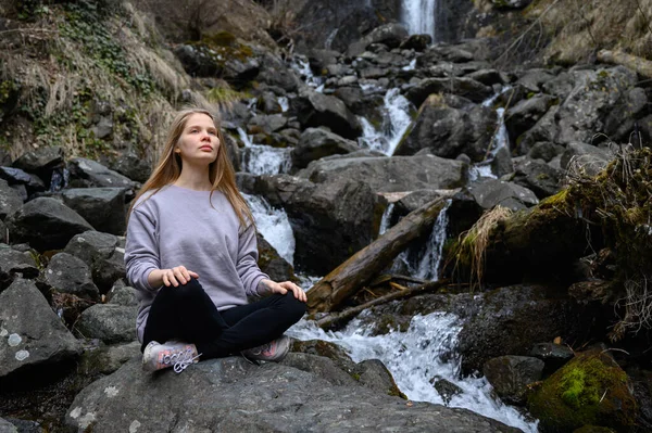 Mädchen sitzt auf einem großen Stein und meditiert in der Nähe eines Wasserfalls neben einem Bergfluss. — Stockfoto