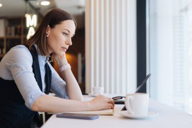 Kafeteryada oturan düşünceli bir kadın ve pencerenin yanında bir kahve.