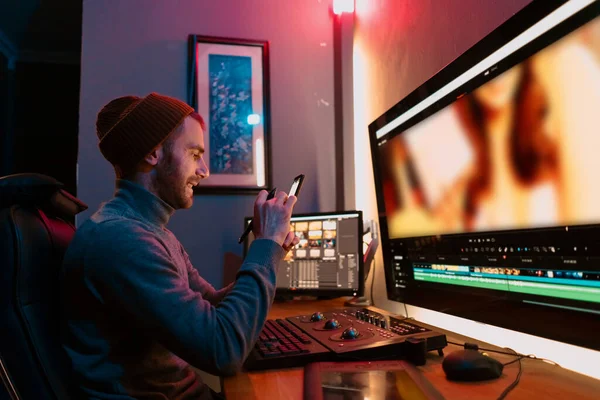 Muž Video Editor sedí ve svém počítači pomocí telefonu — Stock fotografie