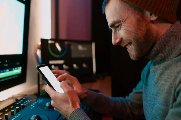 Editor de vídeo masculino sentado en su computadora usando la pantalla en blanco del teléfono —  Fotos de Stock