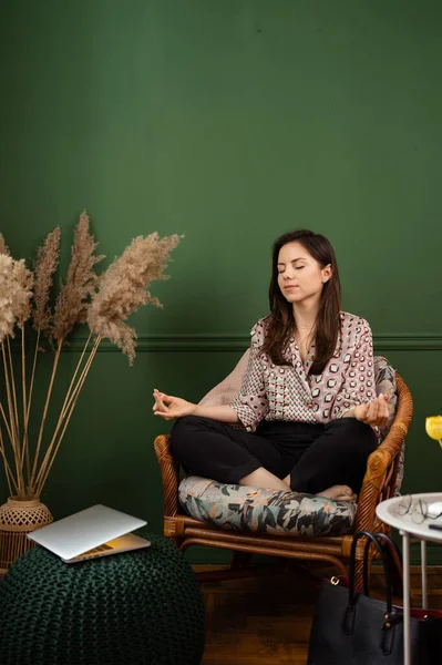 Mujer joven sentada con los ojos cerrados en sillón a laptop en padmasana yoga pose home office. — Foto de Stock