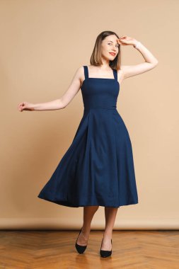 Mavi elbiseli genç kadın bej arka planda poz veriyor.