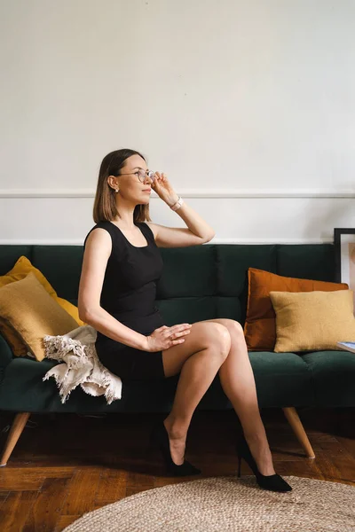 Hermosa mujer en vestido negro y anteojos sentados en un sofá en el interior — Foto de Stock