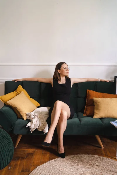 Gyönyörű nő fekete ruhában és szemüvegben ül egy kanapén a házban. — Stock Fotó