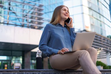 Kadın cep telefonuyla konuşuyor ve basamaklarda otururken laptopuyla çalışıyor.