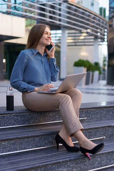 Mujer hablando por teléfono móvil y trabajando con su portátil mientras está sentada en los escalones — Foto de Stock