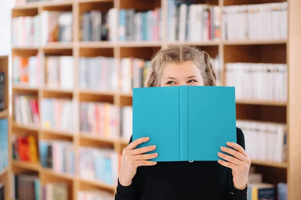 Девушка прячет лицо за книгой с книжной полкой на заднем плане — стоковое фото