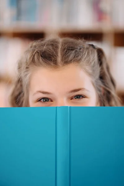 Flicka gömmer ansikte bakom en bok med bokhylla i bakgrunden — Stockfoto