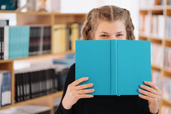 Flicka gömmer ansikte bakom en bok med bokhylla i bakgrunden — Stockfoto