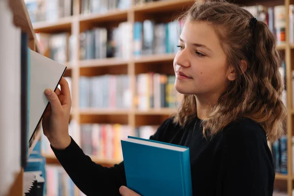 Tonårstjej plockar en bok på en bibliotekshylla — Stockfoto