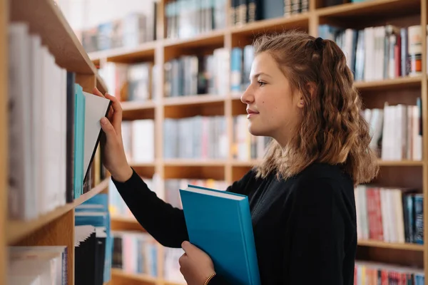 Tonårstjej plockar en bok på en bibliotekshylla — Stockfoto