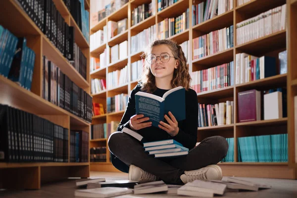 Όμορφη κοπέλα σπουδάζει διαβάζοντας ένα βιβλίο, ενώ κάθεται στο πάτωμα ανάμεσα στα βιβλία στο βιβλιοπωλείο — Φωτογραφία Αρχείου