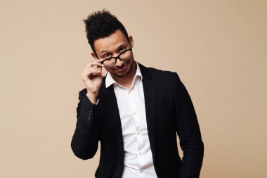 Siyah takım elbiseli, gözlüklü, bej arkaplanda poz veren Afro yakışıklı bir adam.