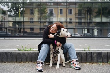 Güzel bir kadın sokak arka planında Dalmaçyalı köpeğe poz veriyor ve sarılıyor.