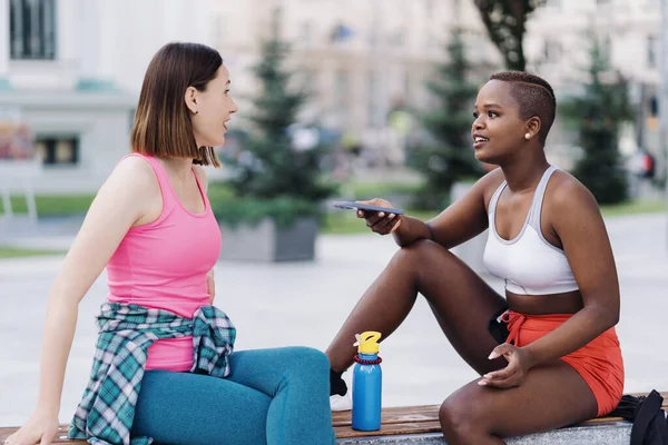 Two multiethnic curvy girls relaxing after exercise session, outdoor communicating. — Stock Photo, Image