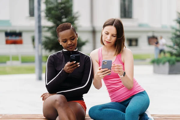 Duas meninas multiétnicas relaxando após sessão de exercício, comunicando ao ar livre e usando smartphone — Fotografia de Stock