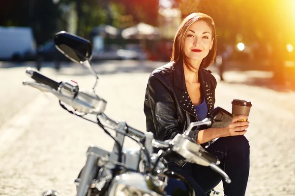 Biker meisje in een lederen jas op een motorfiets drinken koffie — Stockfoto