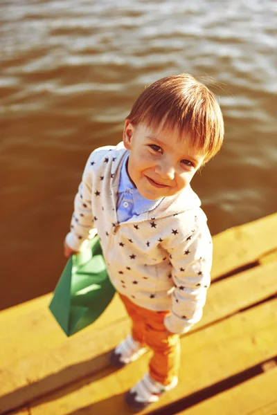 Niño jugando con papel de juguete barco junto al lago — Foto de Stock