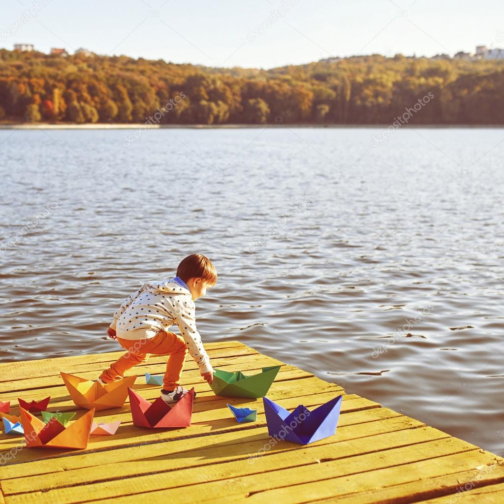 Little boy playing with toy paper ship by the lake