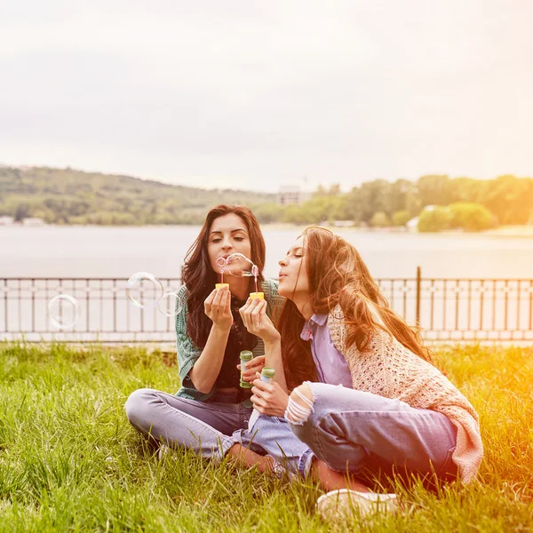 Two cheerful sisters sitting on the grass and blowing bubbles — Stock Photo, Image