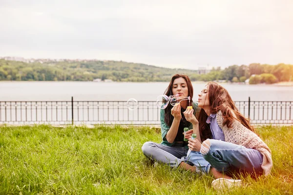 Dvě veselé sestry sedět na trávě a vyfukuje bubliny — Stock fotografie