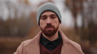 Bereli ve ceketli ciddi bir sakallı adam açık havada kameraya bakıyor..