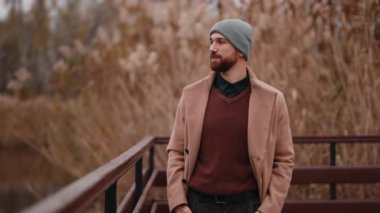 Sakallı ve bereli şık bir adam ahşap bir güvertede duruyor, sonbahar sazlıklarının arasında huzur içinde yansıtıyor ve doğal ortamın tadını çıkarıyor..