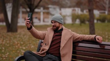 Sakallı bir adam, güzel bir sonbahar gününde mutlu bir şekilde parktaki bankta oturup telefonuyla selfie çekiyor..