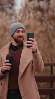 Şık bir adam, açık havada telefonuyla bir video kaydederken sıcak bir içecekten zevk alır. Teknoloji ve sosyal bağlantıyı kucaklar..