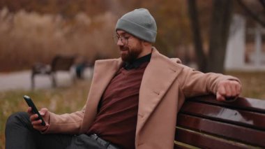 Şık ceketli ve bereli bir adam, sonbaharda parktaki bankta dinlenirken akıllı telefonunu kullanır..