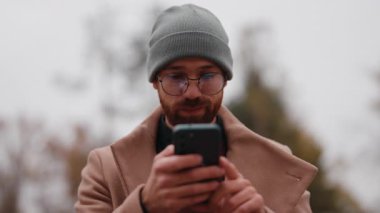 Bereli, gözlüklü ve ceketli şık bir genç adam açık havada cep telefonunu aktif olarak kullanıyor..