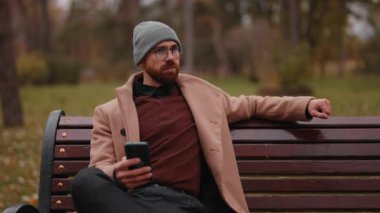 Şık ceketli ve bereli düşünceli genç bir adam cep telefonunu kullanarak bir bankta dinleniyor..