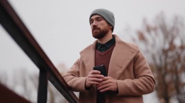 Sıcak bir palto ve bereyle şık bir genç adam serin ve bulutlu bir sonbahar gününde açık havada sıcak bir içecek içiyor..