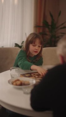 Odaklanmış genç bir kız, rahat bir yuva ortamında bir yetişkinle stratejik bir masa oyunu oynuyor ve nesiller arası bağlantıyı teşvik ediyor..