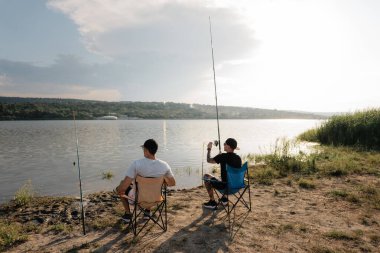 İki arkadaş altın saatinde göl kıyısında balık tutarken dinleniyor, açık havanın tadını çıkarıyorlar, sakin su ve sohbet..
