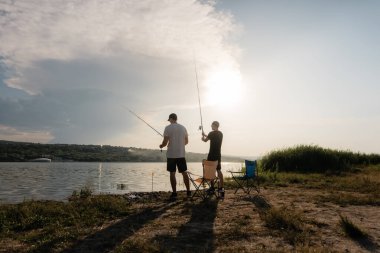 Gün batımında göl kıyısından iki arkadaş, sakin ve yansıtıcı suya doğru çizgiler çiziyorlar..