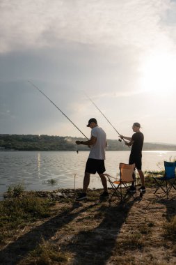 Gün batımında göl kıyısında iki arkadaş olta atıyor ve katlanır sandalyelerde dinleniyor..