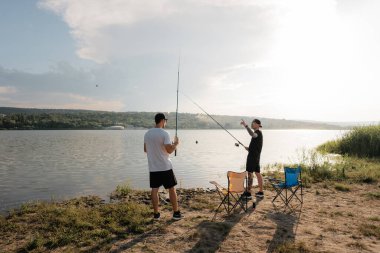 Gün batımında sakin bir gölde balık tutan iki adam, katlanır sandalye ve aletlerin yanında sohbet ediyorlar..