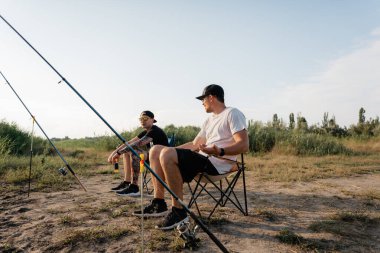 İki genç adam katlanır sandalyelerde oturur, çimli bir nehir kıyısında çubuklarla sohbet eder ve balık tutarlar..