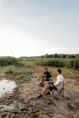 İki arkadaş çimenli bir sahilde katlanan sandalyelerde oturur gün batımında çubuklarla sohbet eder ve balık tutarlar..