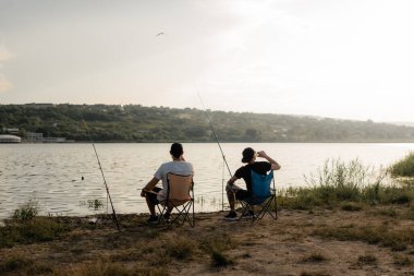 İki arkadaş göl kıyısındaki katlanan sandalyelerde oturur, balık tutar ve huzurlu bir yaz günbatımının tadını çıkarırlar..