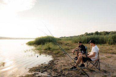 İki adam çimenli bir göl kıyısında iskemlelerin üzerinde oturup gün batımında ellerinde içki ve oltalarla bekliyorlar..
