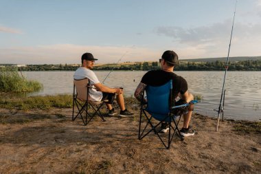 İki adam göl kıyısında katlanan sandalyede dinlenirken balık tutuyor ve yaz akşamı sohbetinin tadını çıkarıyor..