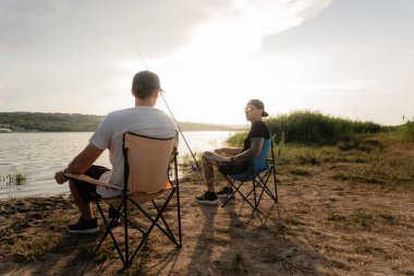 İki arkadaş, sıcak ve rahat bir günbatımı anında, göl kıyısında kamp sandalyelerinde balık tutarlar..