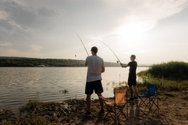 Gün batımında göl kıyısında balık tutan iki arkadaş katlanır sandalyelerin yanında durgun suya halatlar atıyorlar..
