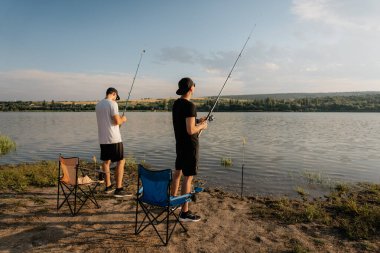İki arkadaş altın saatinde göl kıyısında balık tutuyor, açık havada sakin bir yaz akşamının tadını çıkarıyorlar..