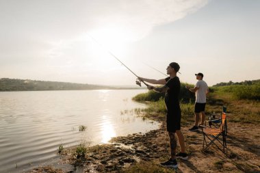 İki arkadaş, altın saatinde sakin bir göl kıyısından, balık tutmak, dinlenmek ve doğanın tadını çıkararak çizgiler çizdiler..